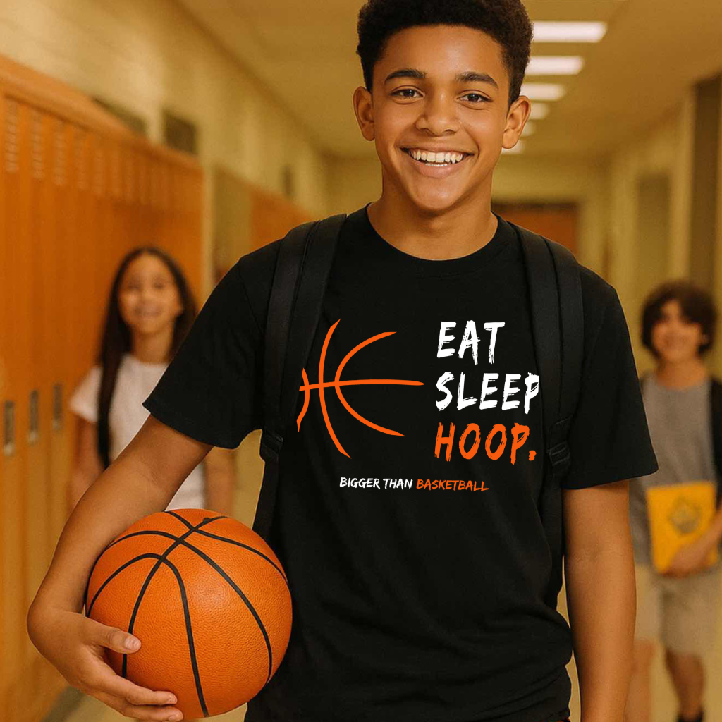Young boy holding a basketball in a school hallway wearing a black t-shirt with a sports-themed design.