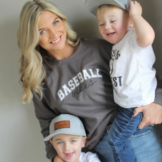 Collection of Woman wearing a 'Baseball Vibes' sweatshirt with two children, one wearing a cap, on a plain background. in a gallery layout