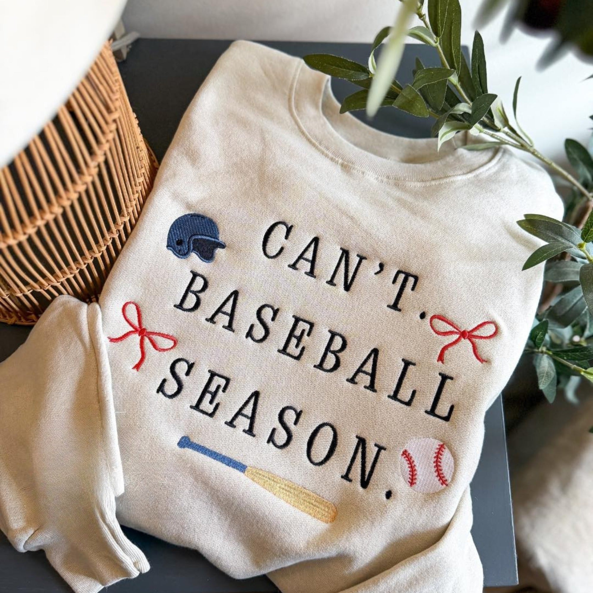 Collection of White sweatshirt with 'Can't. Baseball Season.' text and sports-themed graphics on a surface with a plant in the background. in a gallery layout