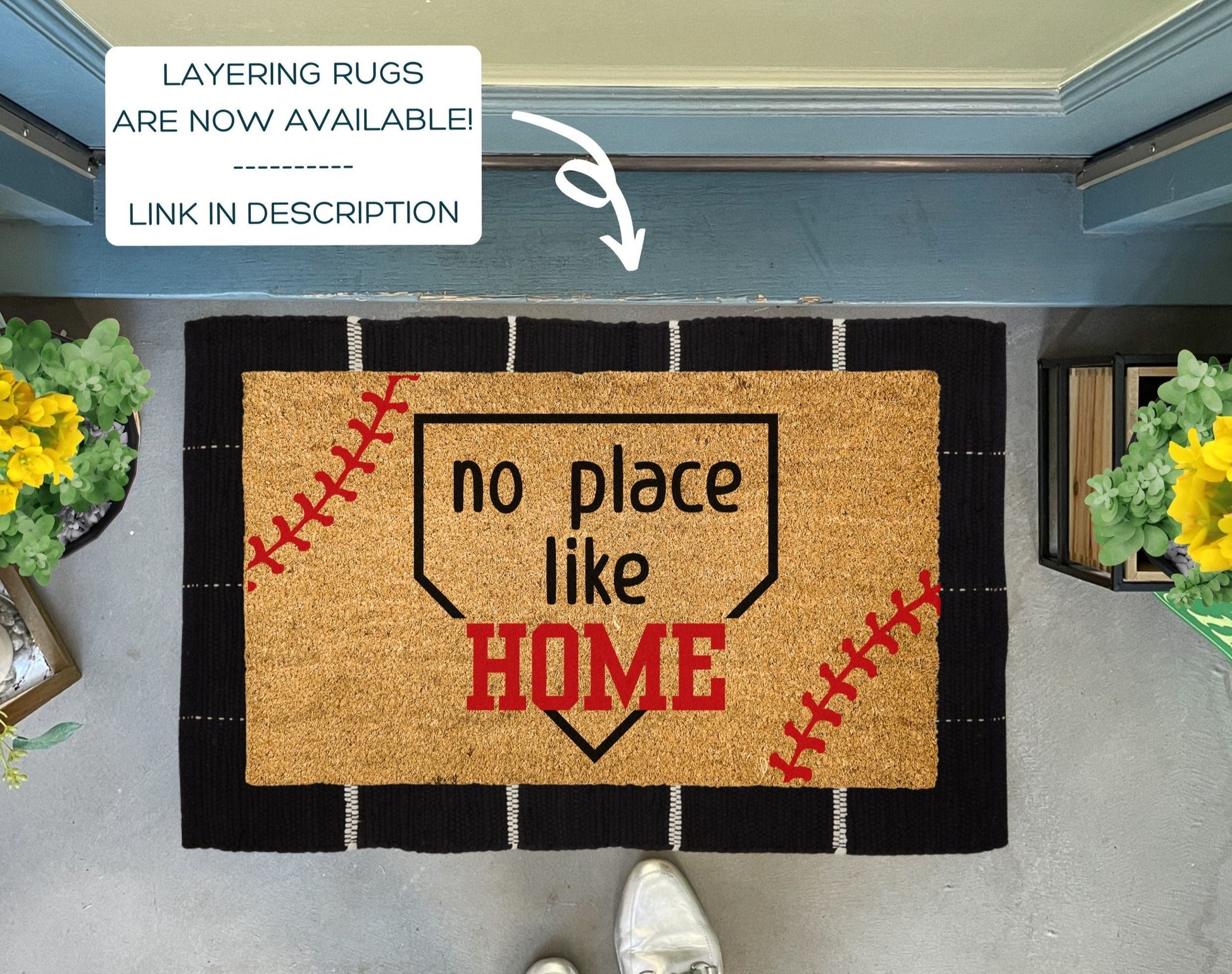 Doormat with 'no place like home' text on a gray floor with plants on either side.