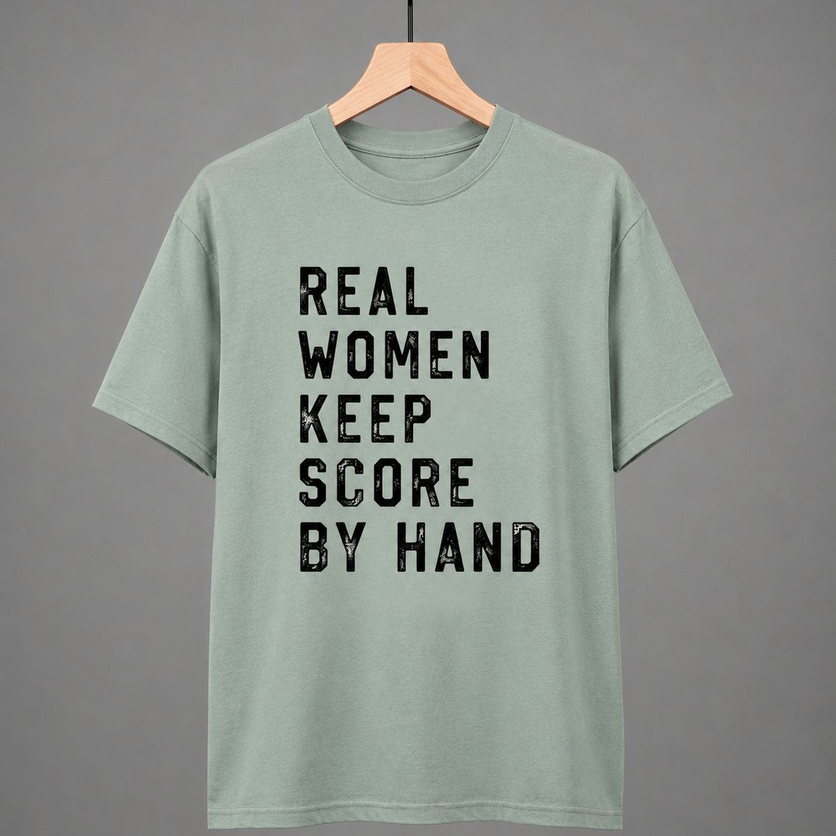 Collection of Green t-shirt with 'Real women keep score by hand' text on a hanger against a gray background in a gallery layout