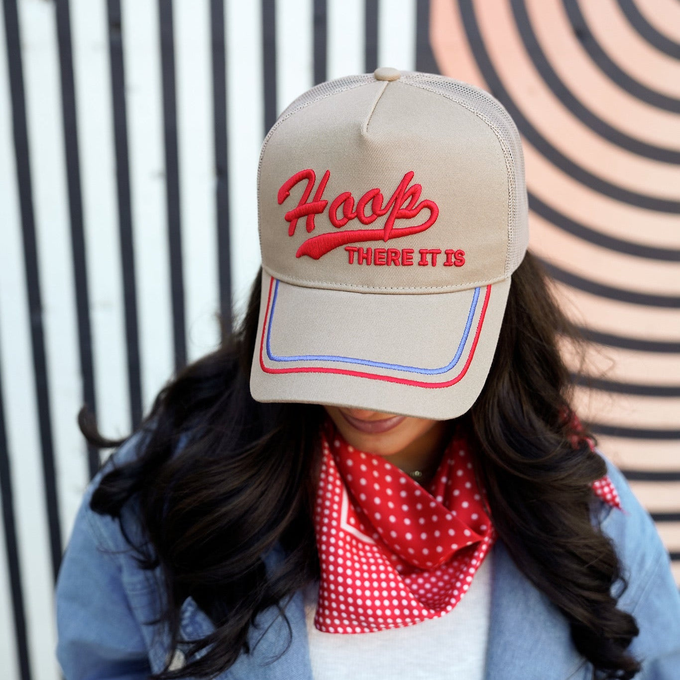 Person wearing a beige cap with red text and a red checkered scarf, standing against a striped background.