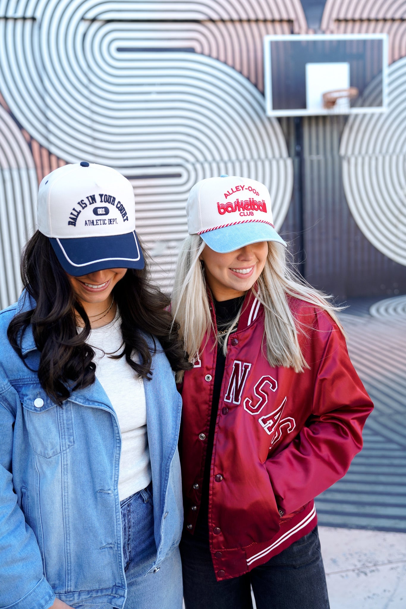 Two women wearing baseball caps and jackets standing in front of a wall with circular patterns.