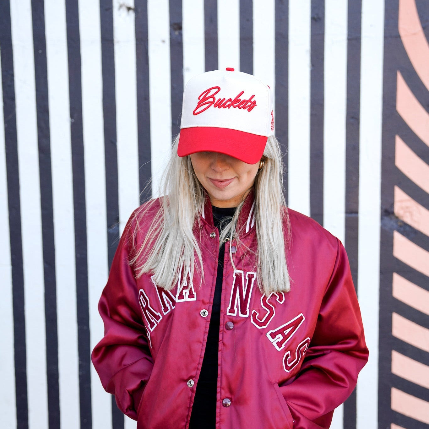 Person wearing a red varsity jacket and white cap with red text against a striped background