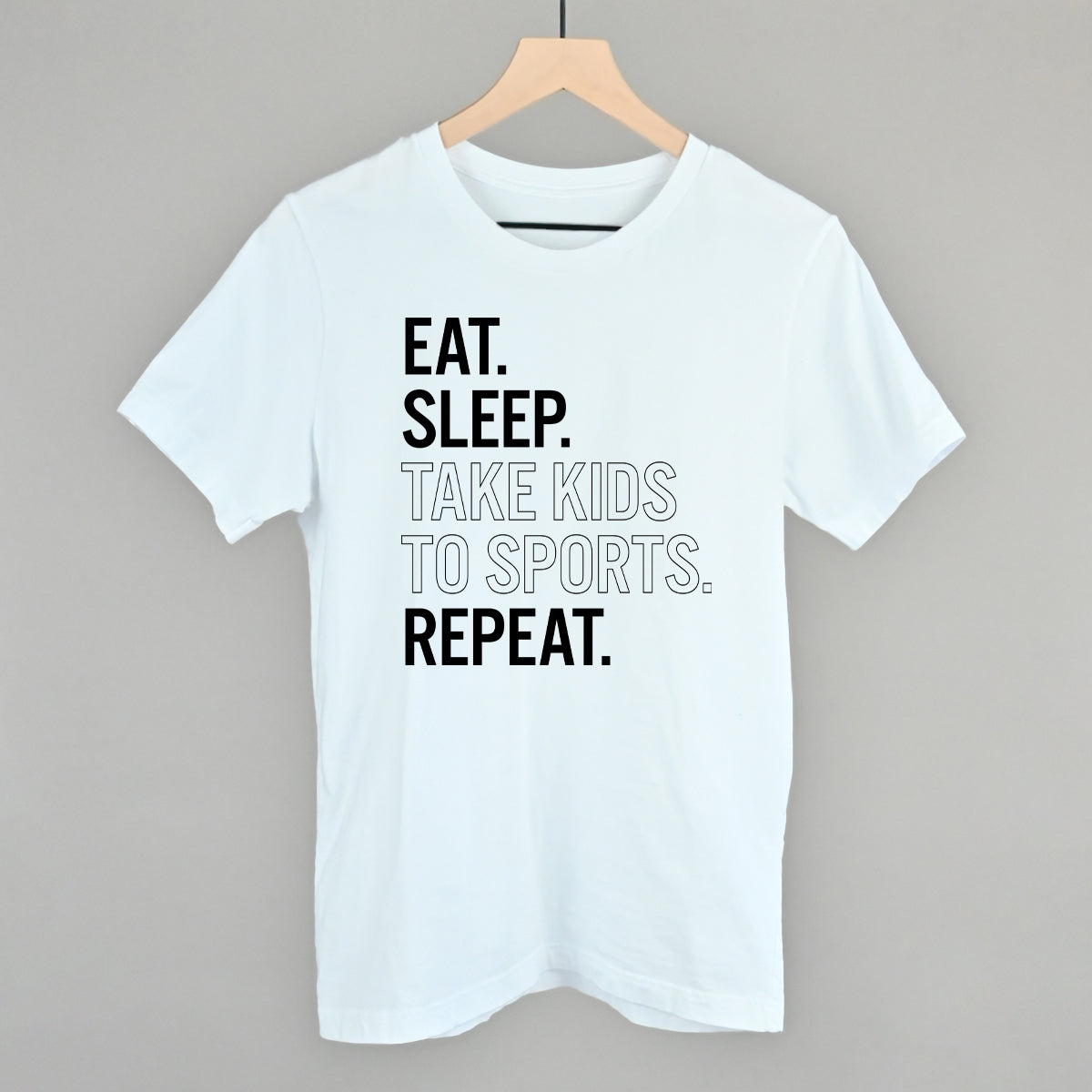 White t-shirt with text 'EAT. SLEEP. TAKE KIDS TO SPORTS. REPEAT.' on a hanger against a gray background