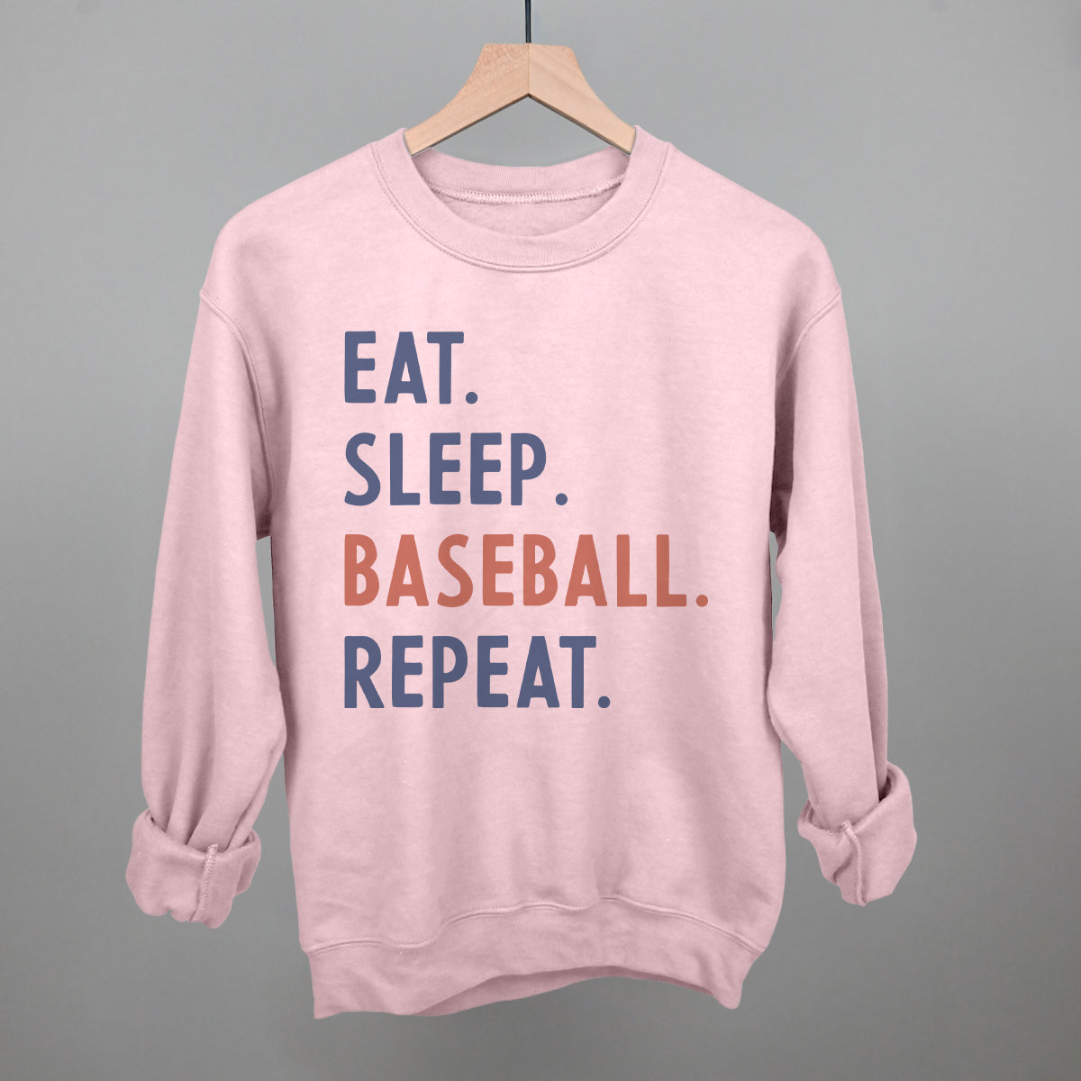 Pink sweatshirt with 'Eat. Sleep. Baseball. Repeat.' text on a hanger against a gray background