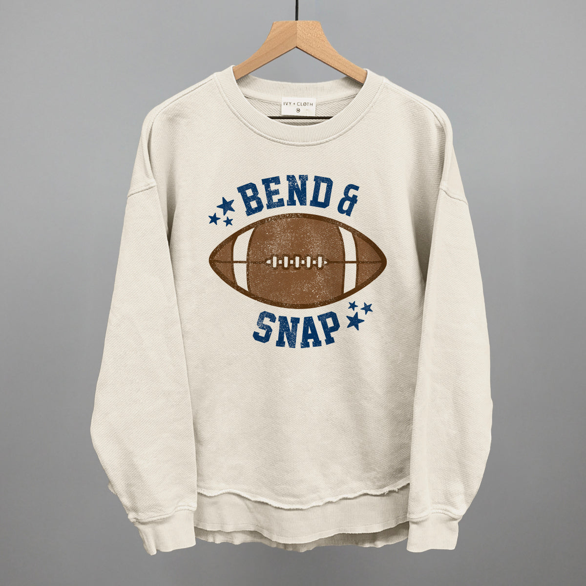 Beige sweatshirt with football graphic and text 'Bend & Snap' on a hanger against a gray background
