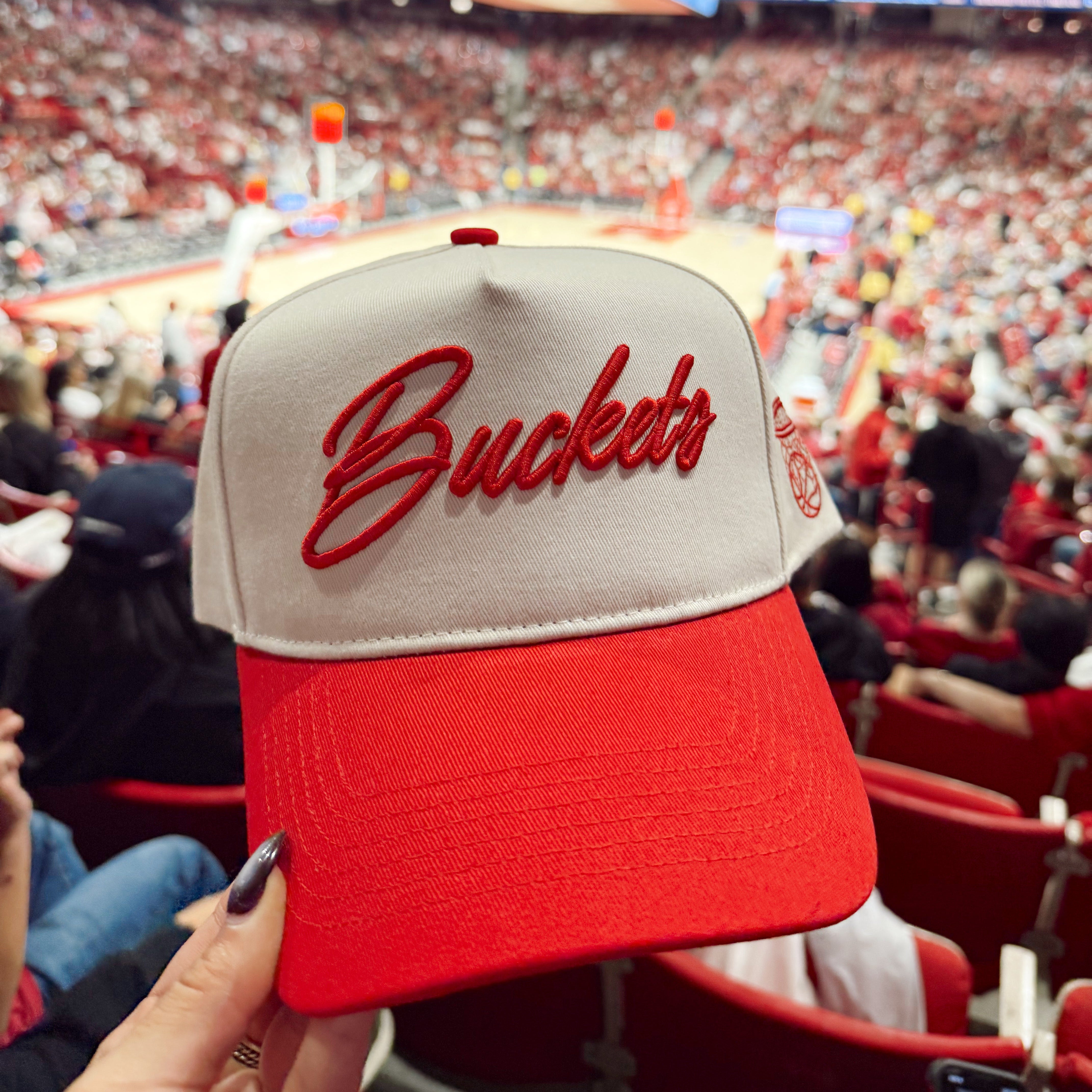 Collection of Red and beige cap with 'Buckets' text held up in front of a sports arena crowd. in a gallery layout