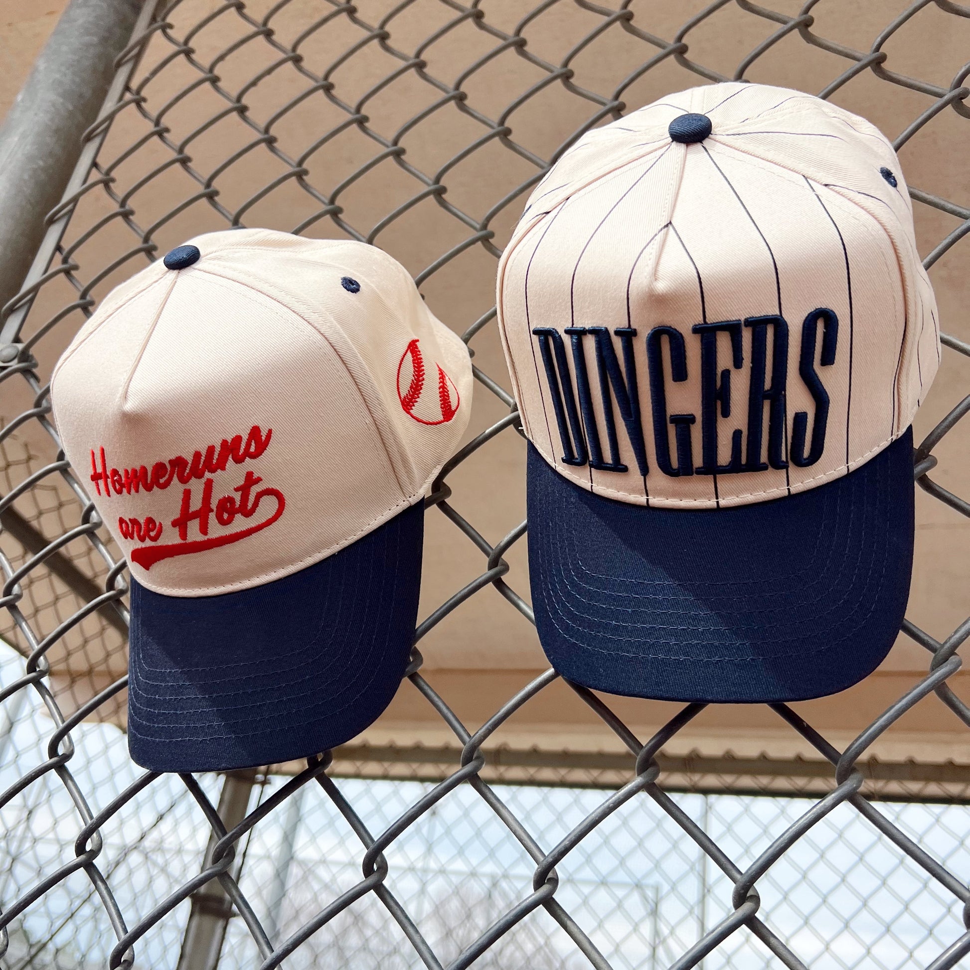Two baseball caps with text on a chain-link fence background