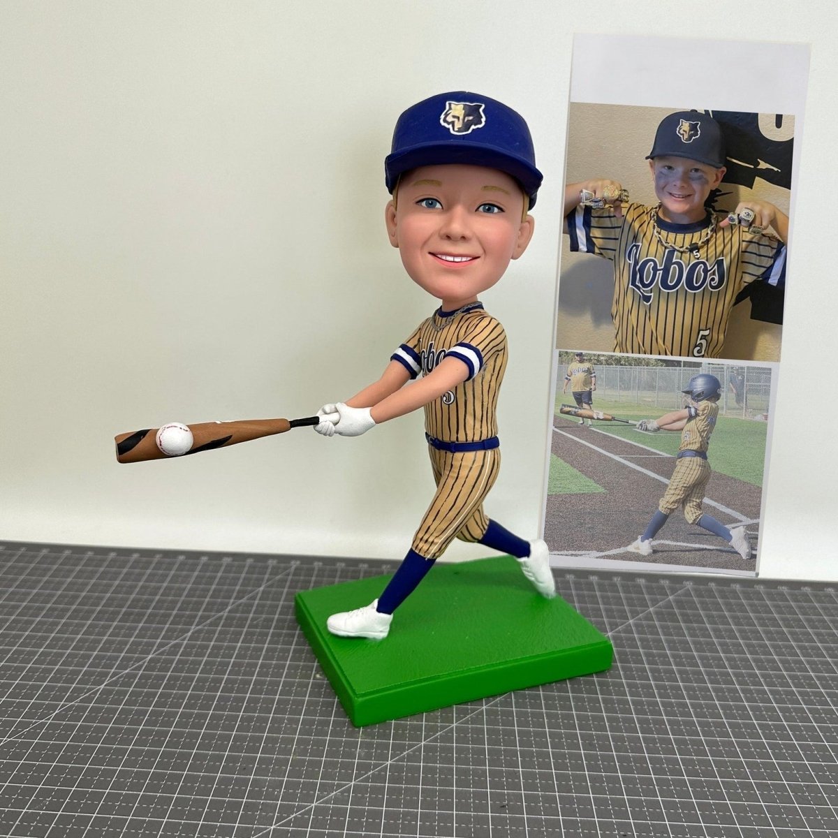 Bobblehead of a baseball player in uniform with a bat, standing on a green base, with a photo of a real-life player in the background.
