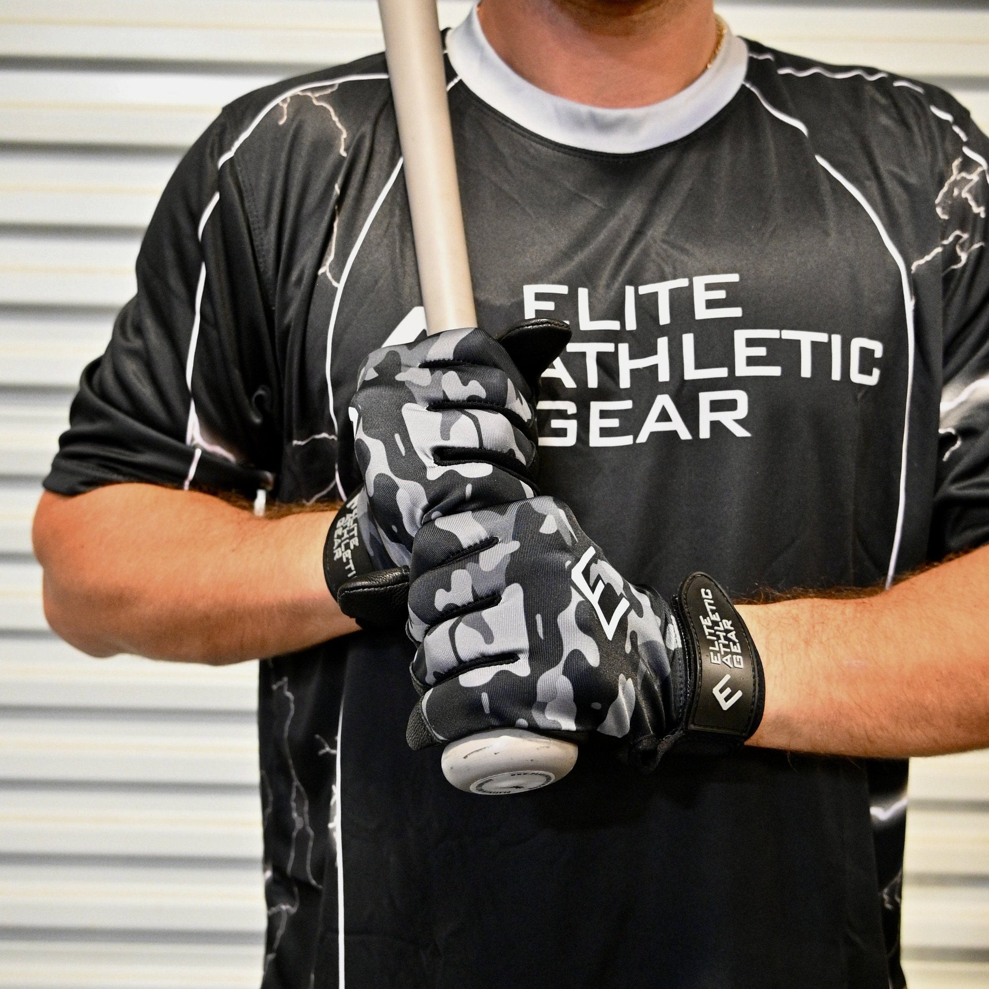 Person wearing a black 'Elite Athletic Gear' shirt and holding a baseball bat with camouflage gloves.