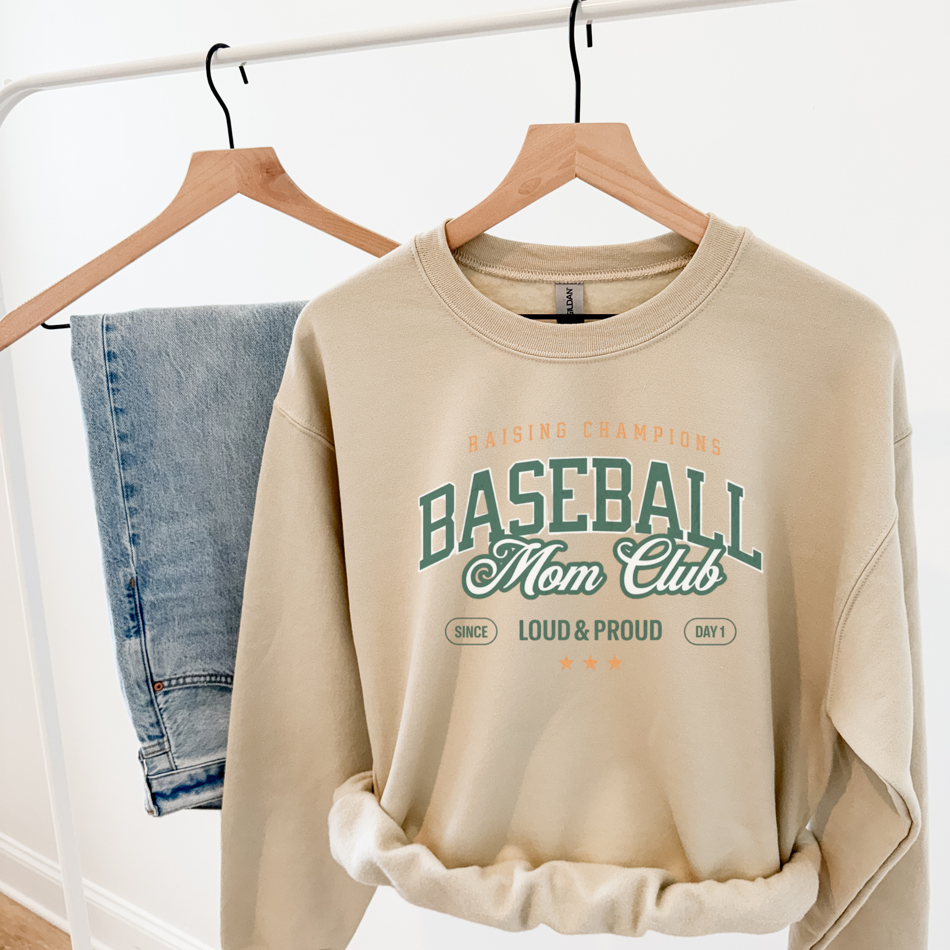 Beige sweatshirt with 'Baseball Mom Club' text on a hanger with jeans in the background.
